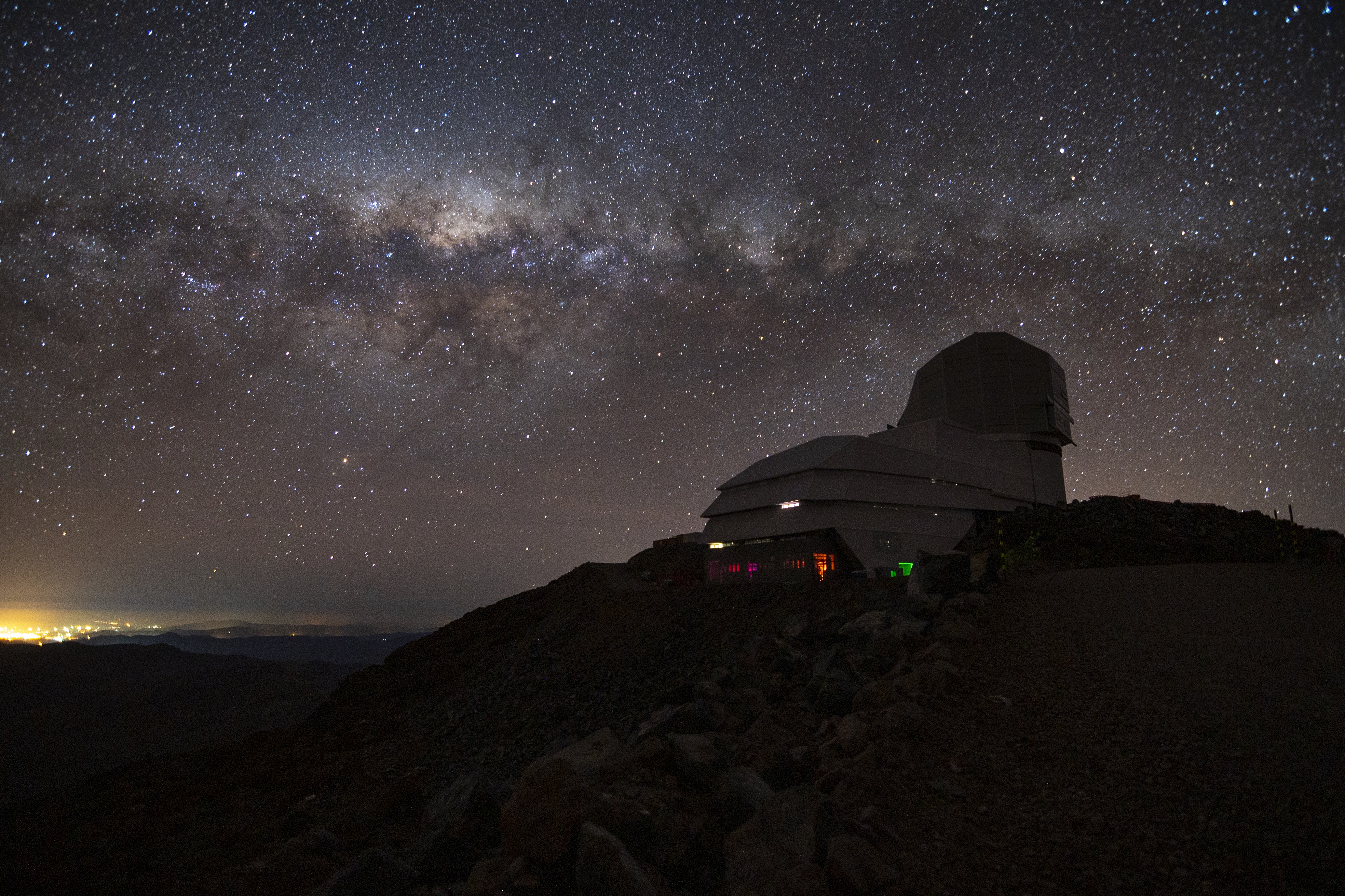 A starry sky illuminates a hillside with a long building on the top. At one end is a square-ish telescope dome, which houses the telescope of the Vera Rubin Observatory.