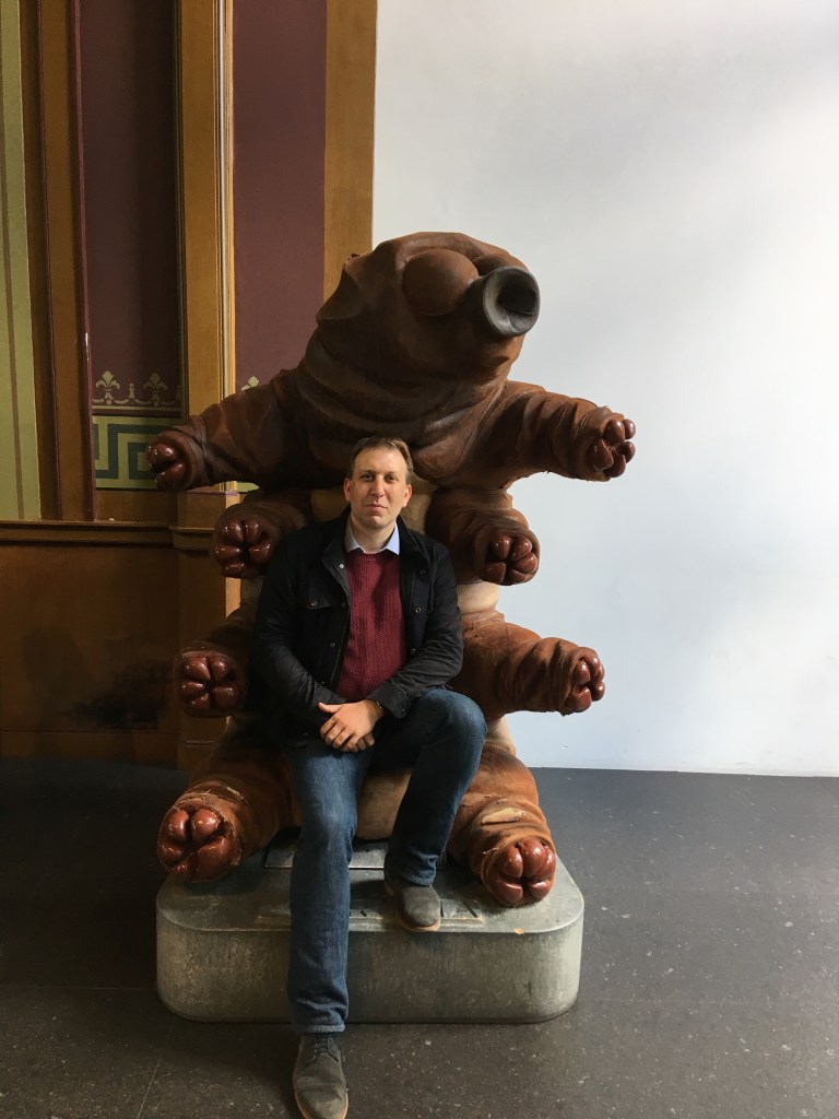 Chris Lintott, wearing a collared shirt, jumper and jeans, poses on a giant tardigrade throne.