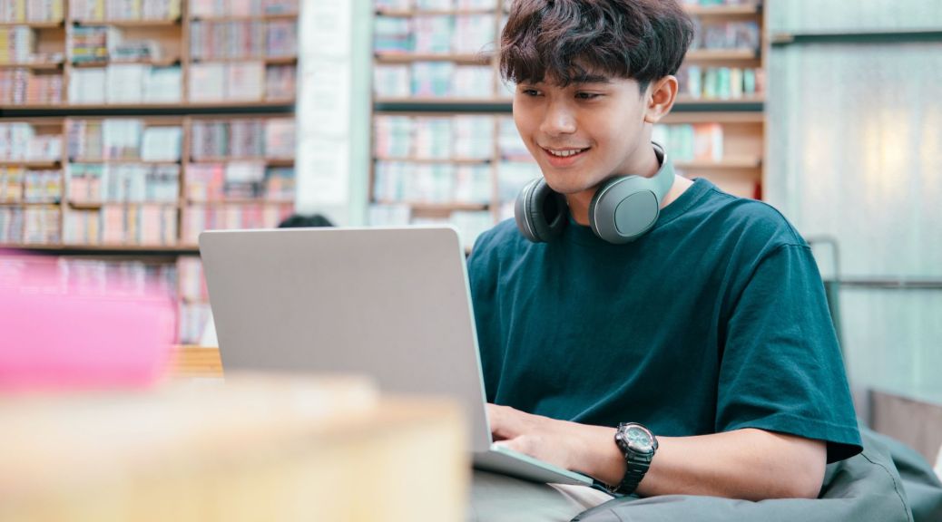 A Young man in a green T-shirt works on his laptop in a library, he looks interested and happy.